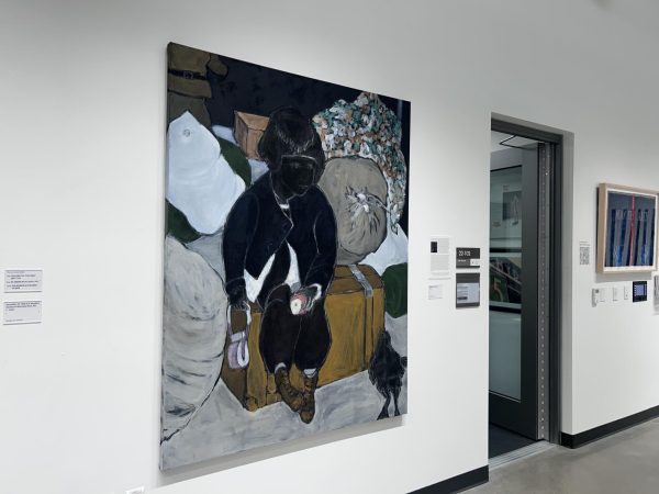 Painting displayed on a white wall in a gallery setting. The painting depicts a dark-skinned figure with undefined facial features, seated on a large brown suitcase surrounded by oversized sacks and a pile of various bundled items. The figure is dressed in dark clothing and boots, and is holding a small object emitting light in their left hand. A black animal is positioned near the figure’s feet. The painting uses muted and dark tones with bold outlines.