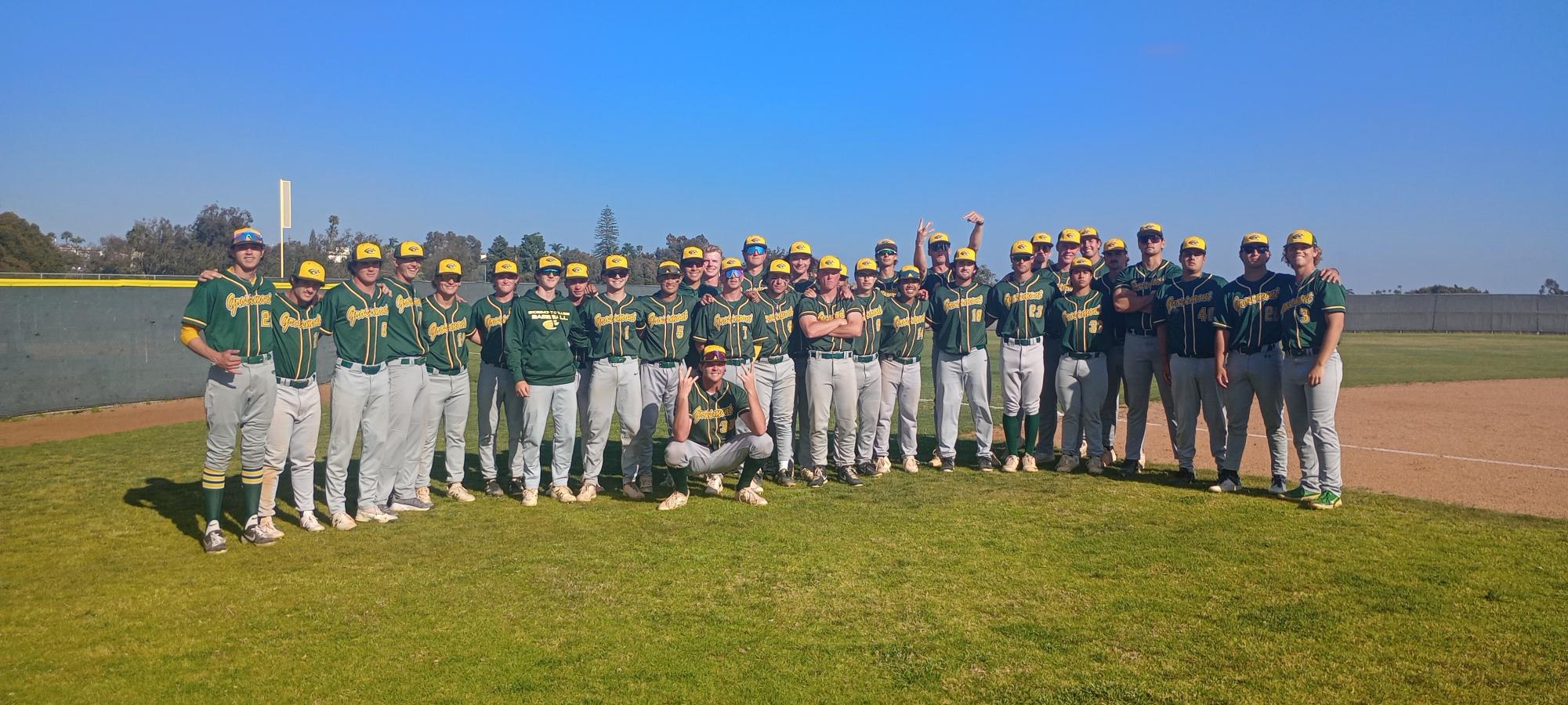 Baseball team in green and yellow uniforms poses together on a sunny field near the infield dirt.