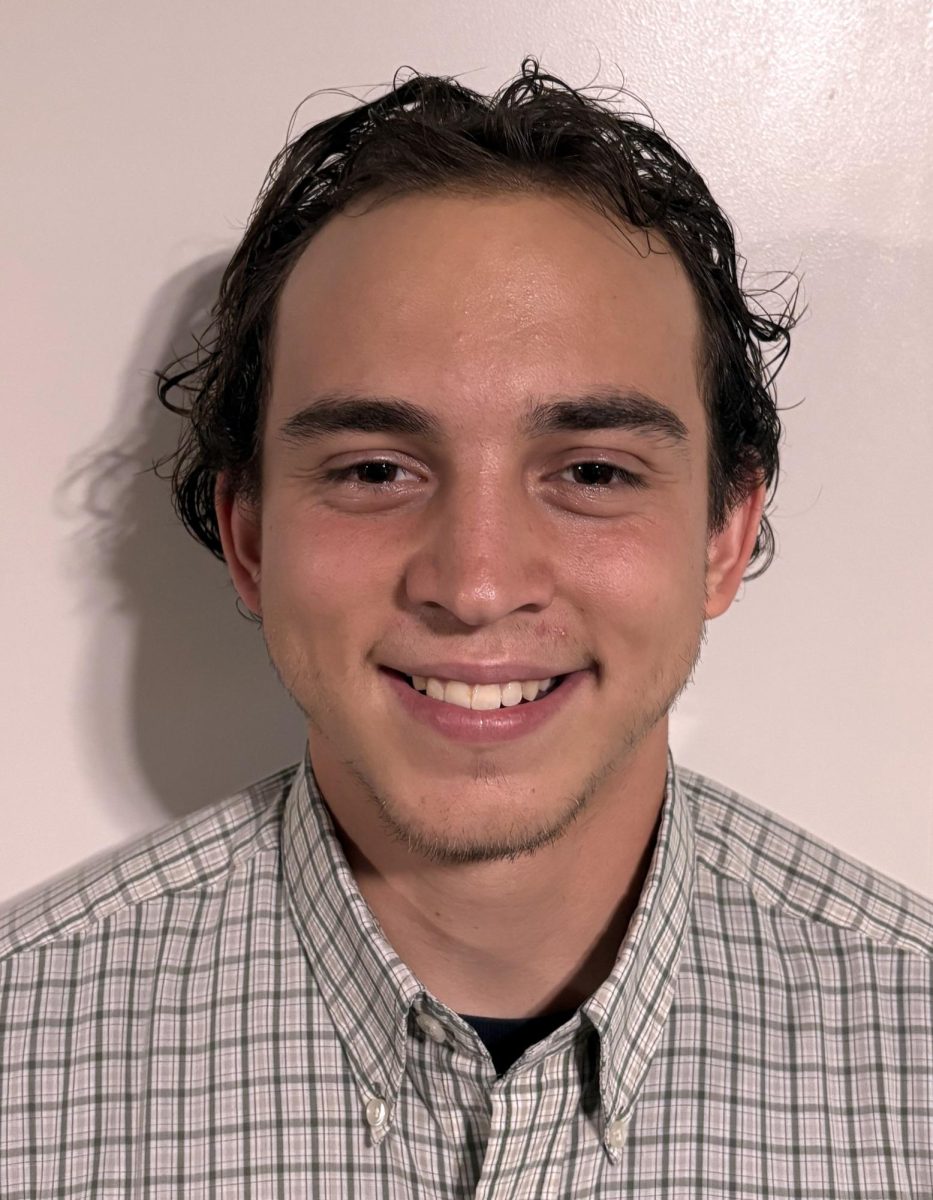 Smiling young man with short curly hair, wearing a plaid button-up shirt, against a plain background.