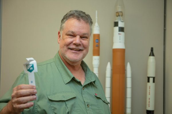 Philip Blanco standing in front of some rocket models in his office and holding one of the paper helicopters he plans to use as an activity in the new PSC-123 course.